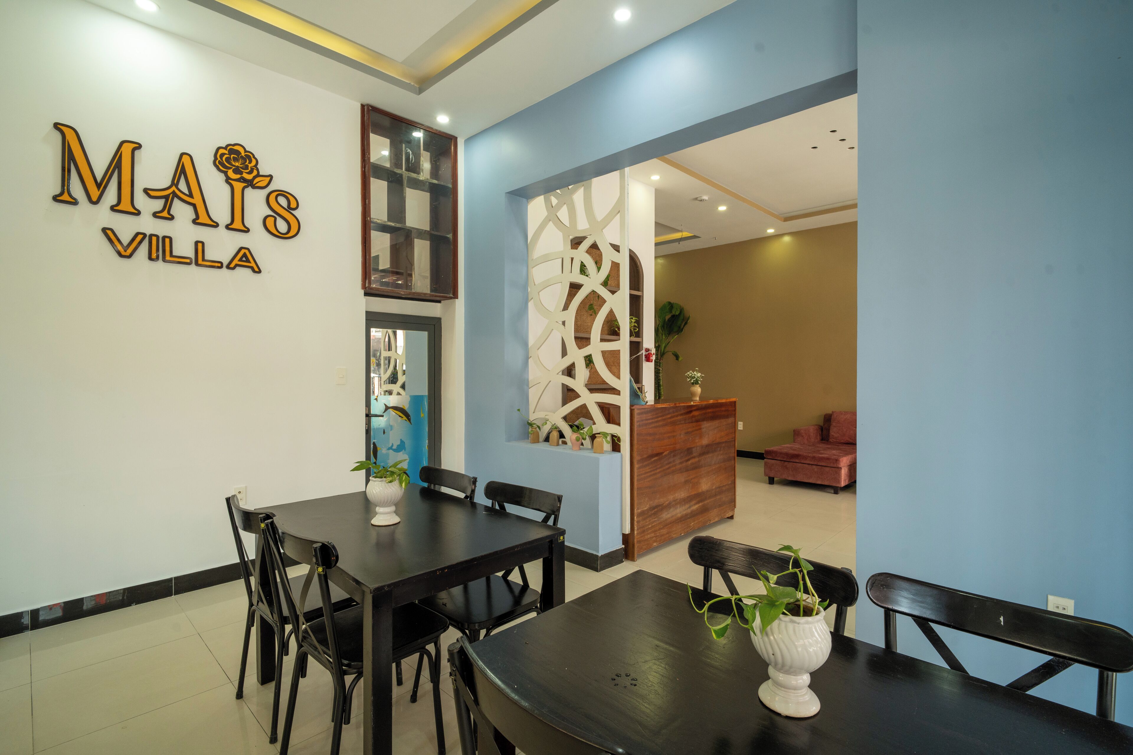 A dining area with a black table and chairs, a reception desk, and a wall with the text "MAIS VILLA".