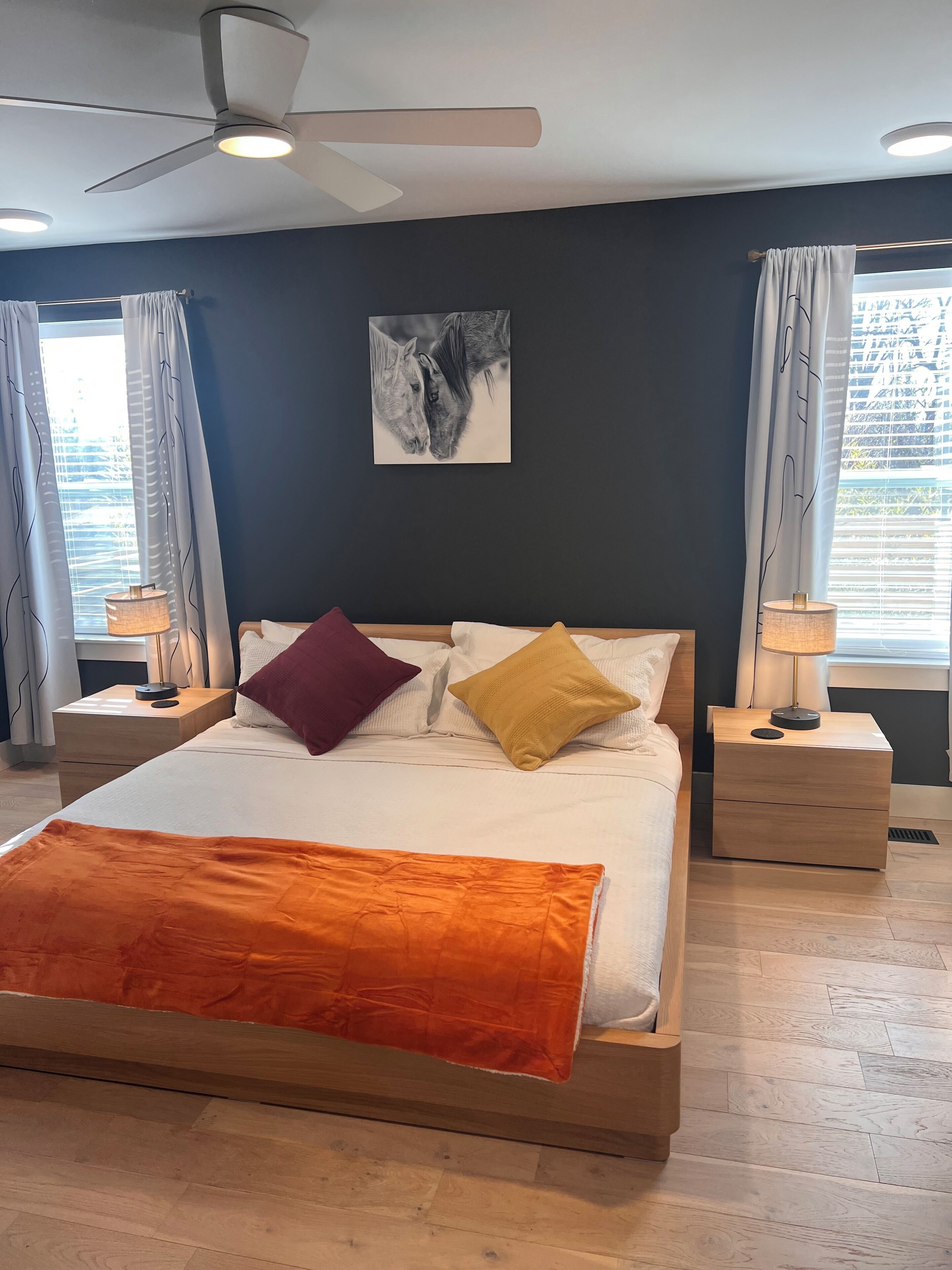Un dormitorio moderno con una cama grande, dos mesitas de noche, un ventilador de techo y una obra de arte colgada en la pared.