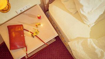 Een net opgemaakt bed met witte lakens, een rode koffer, een klein flesje en een lamp op een nachtkastje.