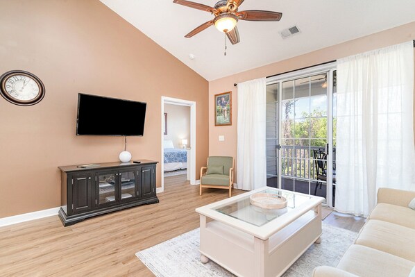 Una sala de estar con televisor, un mueble, una silla, una mesa de centro y una puerta corrediza de vidrio al exterior.