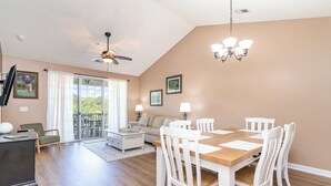 A spacious living room with a wooden dining table, white chairs, a sofa, and a ceiling fan.