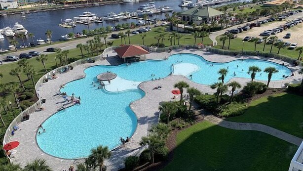 A large, irregularly shaped swimming pool with multiple sections, surrounded by palm trees and a parking area.