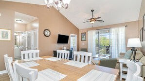 A dining area with a wooden table, white chairs, a ceiling fan, and a TV on the wall.