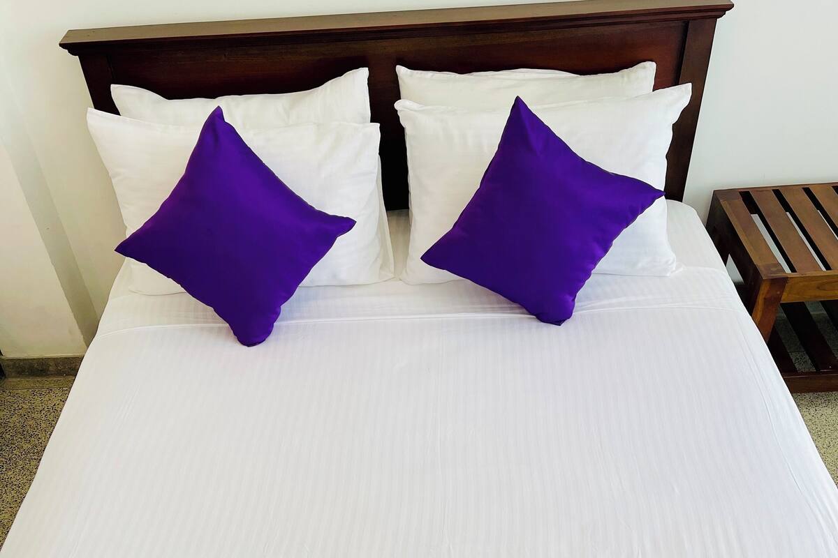 A neatly made bed with white and purple bedding, a wooden headboard, and a small wooden table beside it.