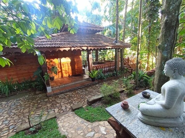 A traditional wooden structure with a tiled roof, surrounded by lush greenery and a stone pathway.