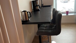 Una cuina moderna amb un taulell fosc, una cafetera i un brollador. Hi ha un tamboret amb peu de crom i seient amb amortidor. Es veu una finestra amb vista a cotxes aparcats.