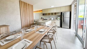 A modern kitchen with a long dining table set for six, equipped with a refrigerator, stove, and ample counter space. - House with private pool in Beira Mar Condominium and private access to the beach. (São Miguel dos Milagres)