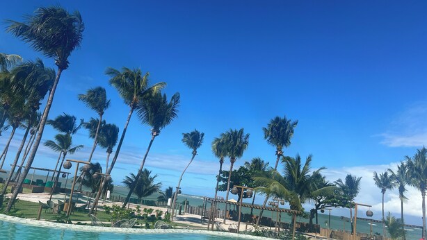 A resort pool area with palm trees, clear blue sky, and a view of the ocean. - House with private pool in Beira Mar Condominium and private access to the beach. (São Miguel dos Milagres)
