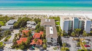 An aerial view of a coastal hotel with multiple buildings, a beach in the background, and a clear blue sky.