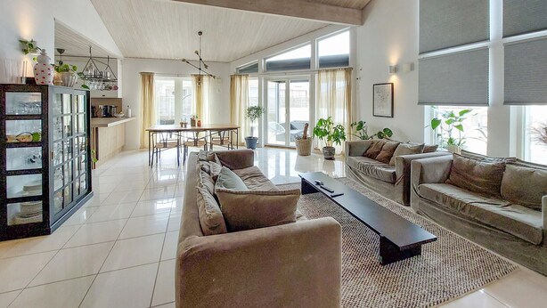 A spacious living area with a large sofa set, a coffee table, and a dining table. There are potted plants and a large window providing natural light.