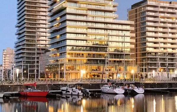 A modern high-rise building with multiple balconies and large windows, situated next to a calm waterfront with several boats docked.