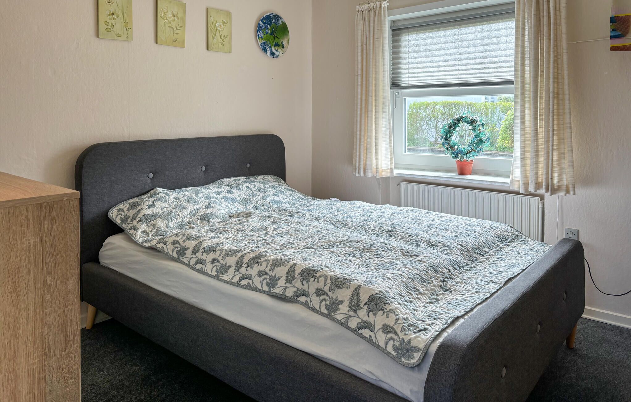 Et soveværelse med en seng, et vindue med gardiner, en radiator og en væg med blomsterudsmykning.