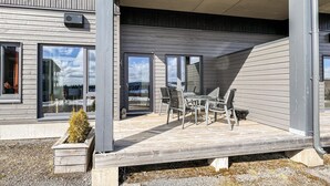 Un patio moderno al aire libre con mesa y sillas de vidrio, rodeado de una terraza de madera y grandes ventanales que ofrecen vistas panorámicas.