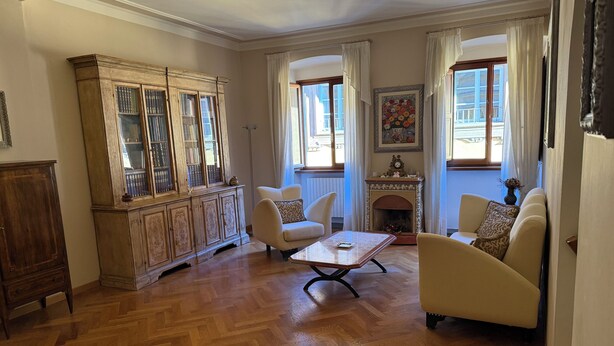 Un salón amplio con suelo de madera en espiga, un mueble de madera grande, dos sillones blancos, una mesita y una chimenea.