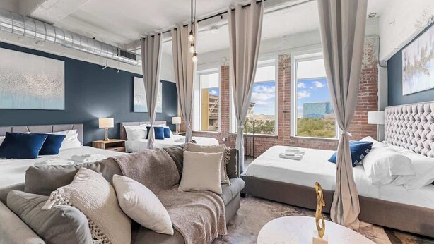 A hotel room with a large bed, a sofa, and a dining table. There are curtains, a ceiling fan, and a view of the city.