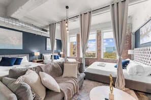 Habitación de hotel con una cama grande, un sofá y una mesa de comedor. Hay cortinas, un ventilador de techo y vistas de la ciudad.