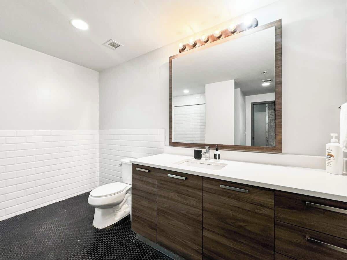 Une salle de bain moderne avec des toilettes blanches, un grand miroir avec éclairage intégré, un lavabo blanc et des meubles en bois foncé.
