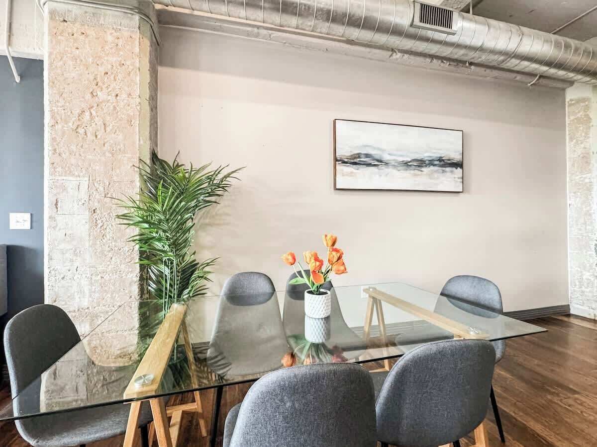 A conference room with a glass table, four grey chairs, a potted plant, and a painting on the wall.