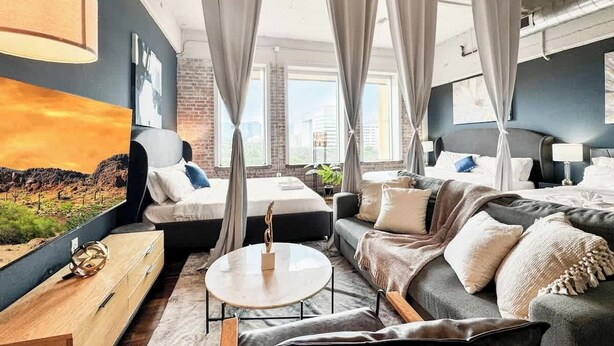 A modern hotel room with a large bed, a sofa, a small table, and a view of the city through large windows.