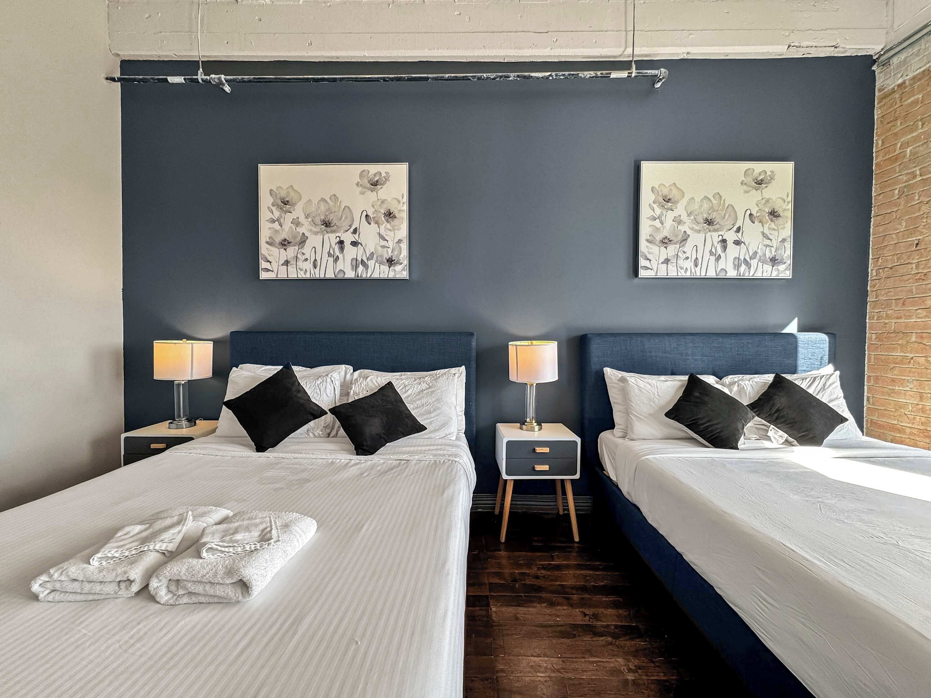 A hotel room with two beds, dark blue headboards, white bedding, black and white floral artwork on the walls, and a small wooden nightstand with a lamp between the beds.