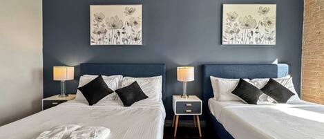 A hotel room with two beds, dark blue headboards, white bedding, black and white floral artwork on the walls, and a small wooden nightstand with a lamp between the beds.