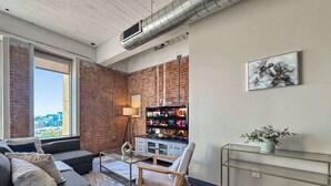 Ein modernes Wohnzimmer mit grauem Sofa, einem Holzstuhl, einem Glastisch und einem Fernseher an einer Ziegelwand.