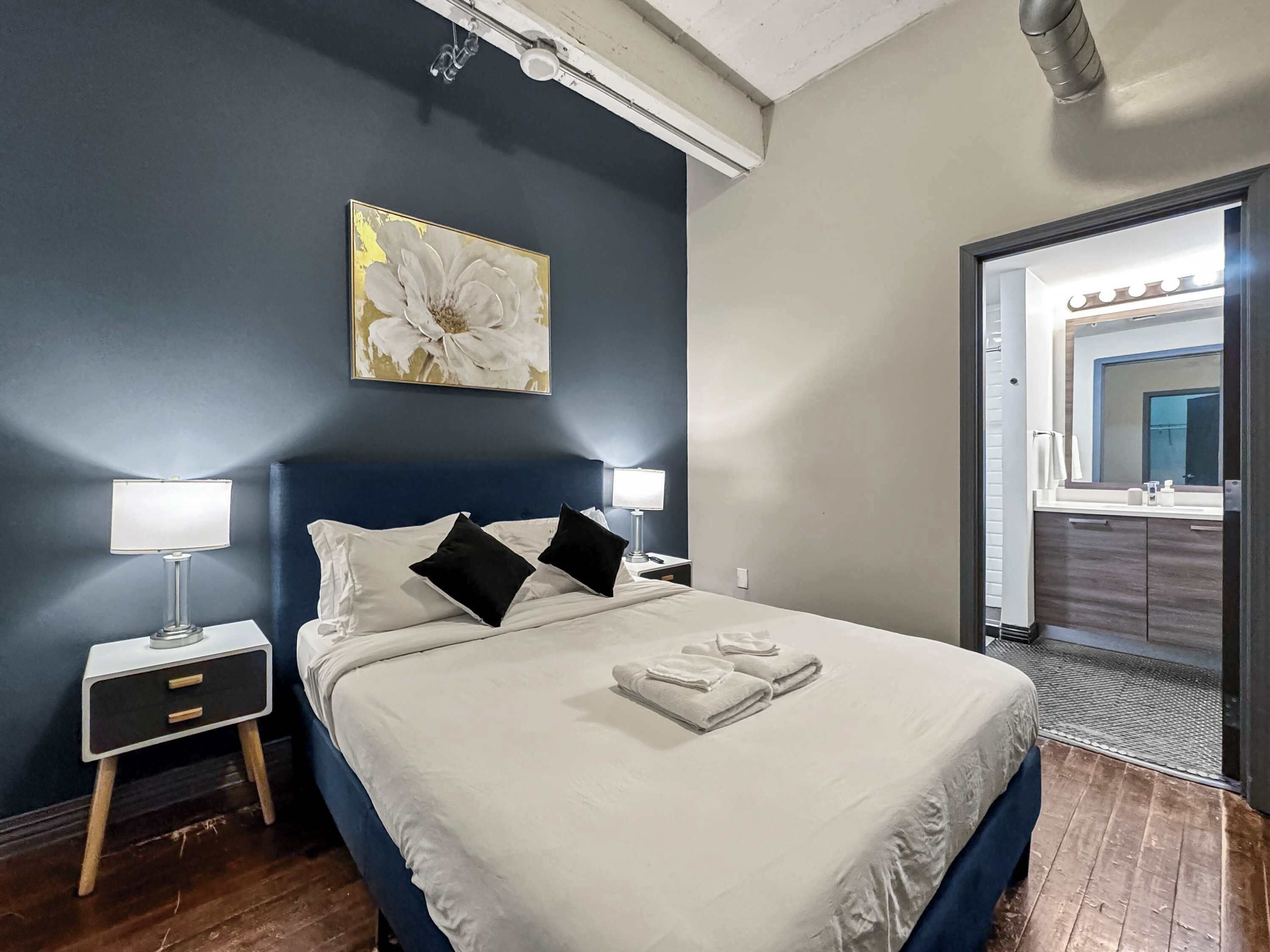 Cama bem arrumada com lençóis brancos e duas almofadas pretas, mesa de cabeceira com abajur, quadro pendurado na parede e banheiro visível por uma porta aberta.