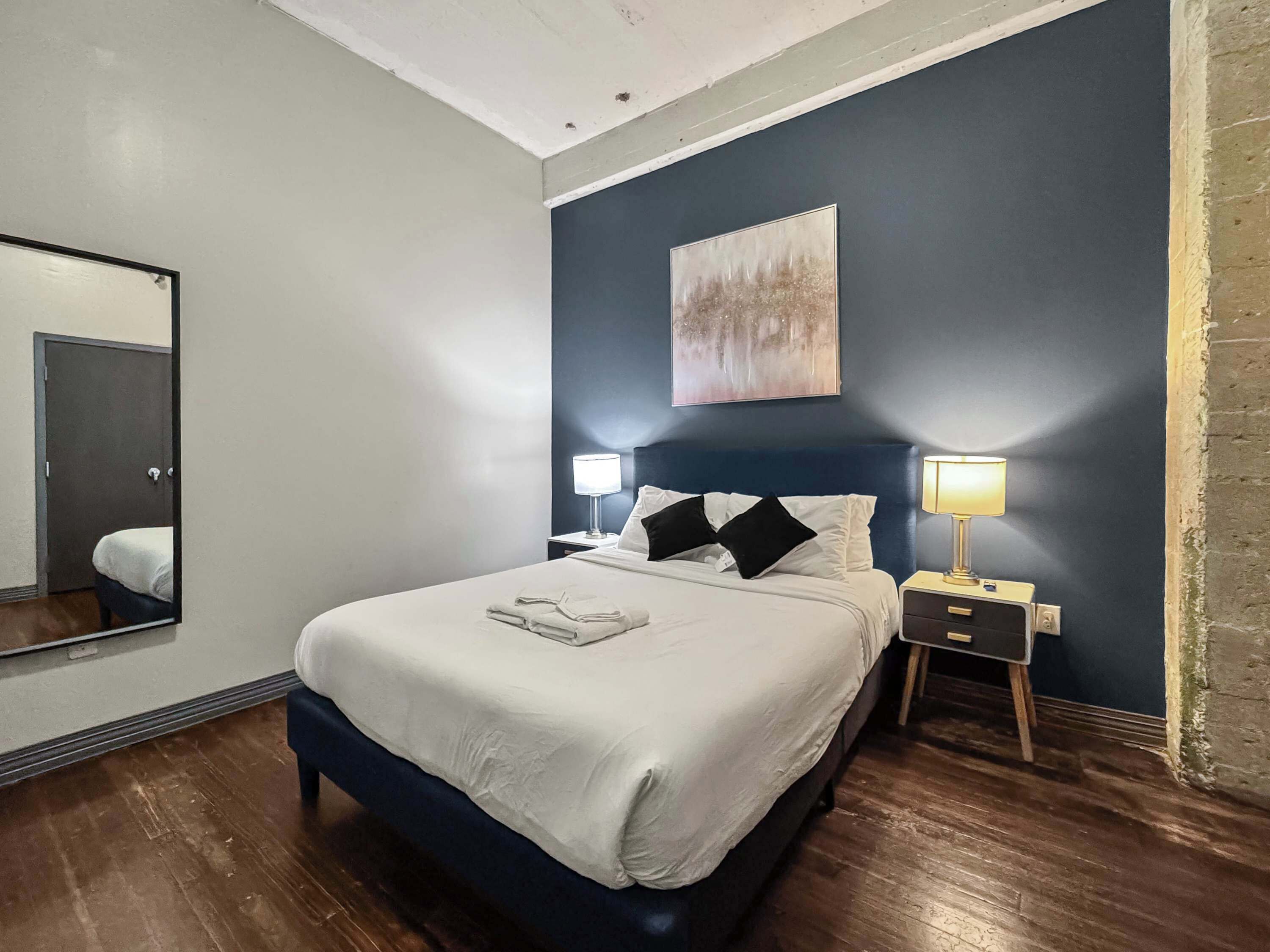 A neatly made bed with white linens and black pillows, a bedside table with a lamp, a large mirror on the wall, and a painting above the bed.