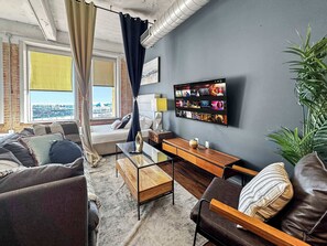 Ein modernes Wohnzimmer mit einer Couch, einem Couchtisch, einem an der Wand befestigten Fernseher und einem großen Fenster mit Vorhängen.