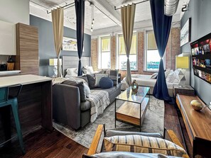 A modern living room with a sofa, armchair, and coffee table. There is a kitchen area with a counter and cabinets. The room has large windows with curtains.