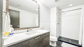 A modern bathroom with a white sink, dark wooden cabinets, a large mirror, and a shower area with white tiles. - Elegant 2BD | DT Dallas | Sleeps 5 | ValetParking (Dallas)