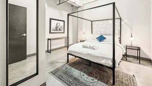 A modern bedroom with a four-poster bed, a framed picture on the wall, and a small bedside table with a lamp. - Elegant 2BD | DT Dallas | Sleeps 5 | ValetParking (Dallas)