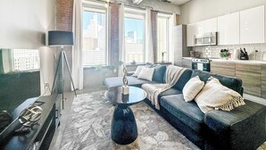 A modern living room with a sofa, coffee table, and a TV. There is a kitchen area with cabinets and appliances. - Elegant 2BD | DT Dallas | Sleeps 5 | ValetParking (Dallas)