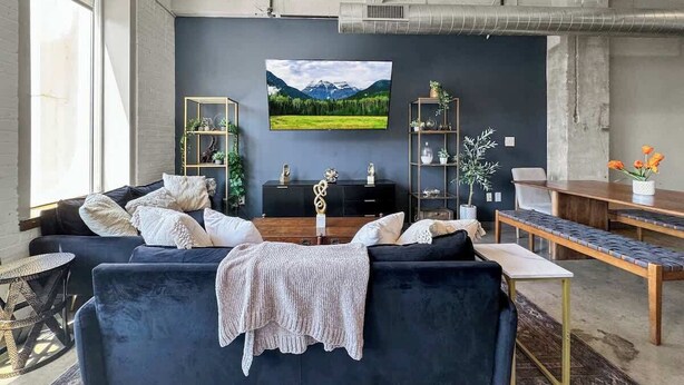 Una sala moderna con un sofá azul oscuro, una mesa de centro y un televisor montado en la pared. Se observa una estantería con objetos decorativos y una planta al lado derecho.