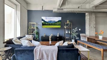 A modern living room with a dark blue sofa, a coffee table, and a TV mounted on the wall. There is a bookshelf with decorative items and a plant on the right side.