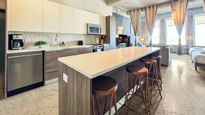 A modern kitchen with a central island, bar stools, and a dining area with a sofa and lamp. - Luxury 2BD | DT Dallas | Sleeps 14 | ValetParking (Dallas)