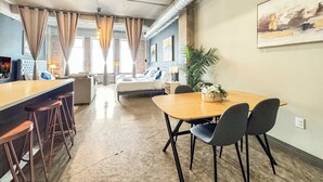 A modern hotel room with a large bed, a sitting area with a sofa and a coffee table, a dining table with chairs, and a large window with curtains. - Luxury 2BD | DT Dallas | Sleeps 14 | ValetParking (Dallas)