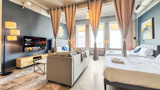 A modern hotel room with a large bed, a sofa, a TV, and a view of the city through large windows. - Luxury 2BD | DT Dallas | Sleeps 14 | ValetParking (Dallas)
