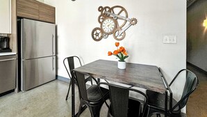 Una cocina con un refrigerador de acero inoxidable, una mesa de comedor de madera con cuatro sillas y un reloj de pared con engranajes.