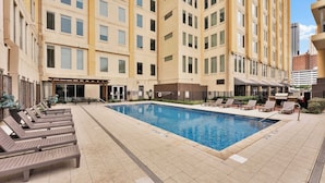 A hotel pool area with sun loungers, a barbecue, and a covered seating area. - Posh 2BR Suite | Texas Core | Fits 6 | Vallet Park (Dallas)