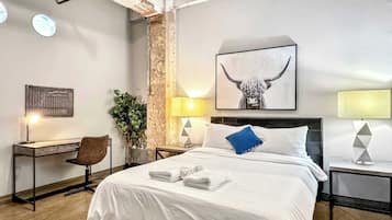 A hotel room with a large bed, a desk, a chair, a potted plant, and a wall-mounted artwork.