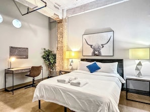 A hotel room with a large bed, a desk, a chair, a potted plant, and a wall-mounted artwork. - Posh 2BR Suite | Texas Core | Fits 6 | Vallet Park (Dallas)