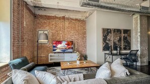 A living room with a brick wall, a television, a coffee table, and a sofa. - Elegant 3BD | DT Dallas | Sleeps 9 | ValetParking (Dallas)