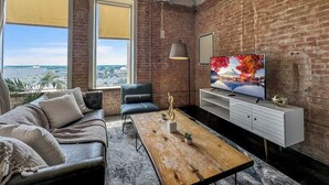 A modern living room with a leather sofa, a wooden coffee table, and a TV on a brick wall. - Elegant 3BD | DT Dallas | Sleeps 9 | ValetParking (Dallas)