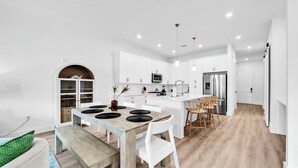 Una cocina moderna con armarios blancos, una isla central y una zona de comedor con mesa de madera y sillas blancas.