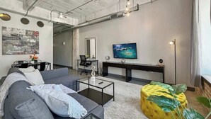 A modern living room with a gray sofa, a glass coffee table, a yellow ottoman, and a flat-screen TV on the wall.