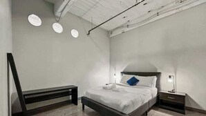 A bedroom with a bed, bedside tables, a bench, and a ceiling with exposed pipes.