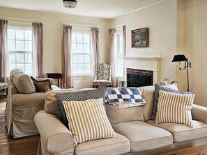 Een woonkamer met een beigekleurige zithoek, een gestreept fauteuil, een open haard en een kroonluchter.