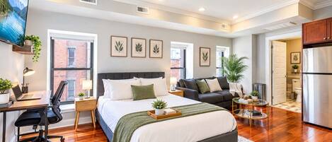 Un dormitorio moderno con cama, escritorio y zona de estar, con ventanas amplias y ventilador de techo.