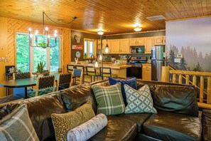 A cozy cabin interior with a leather sofa, dining area, kitchen, and a large window with a view of trees.